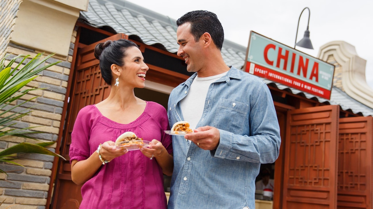 Casal segura pratos de comida em frente ao quiosque da China durante o EPCOT Food & Wine Festival.