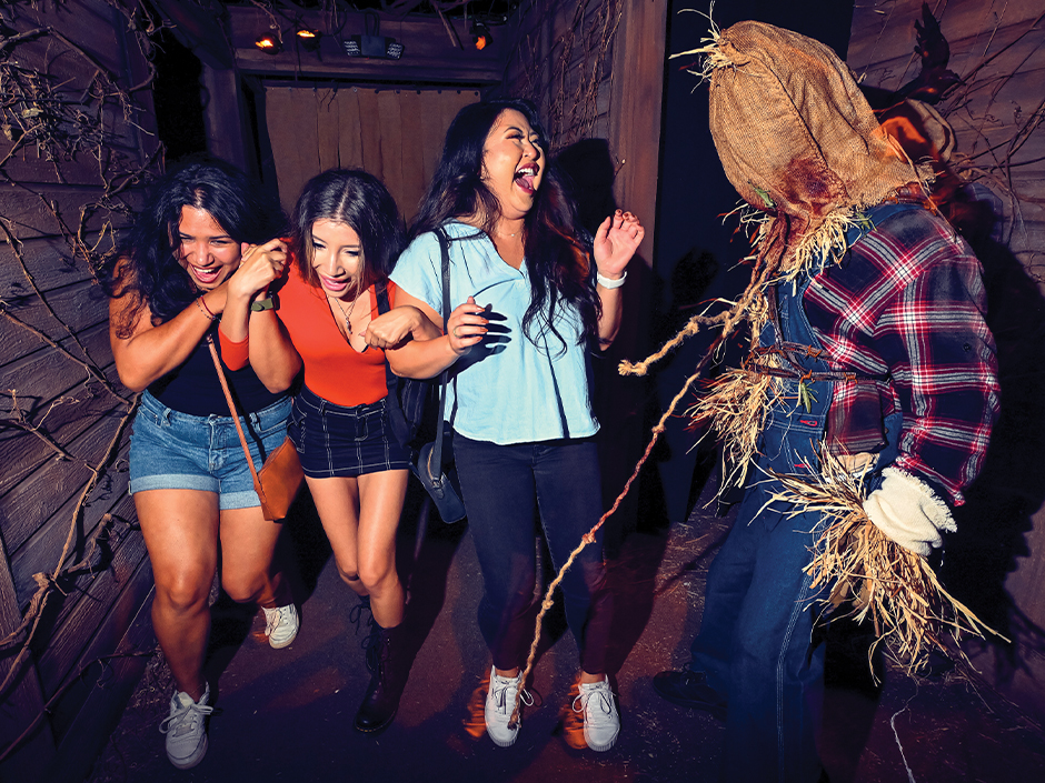 Três visitantes reagem a um scareactor vestido de espantalho dentro de uma casa do Halloween Horror Nights.