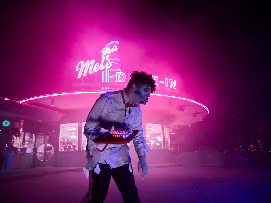 Atendente zumbi aparece entre luzes e fumaça diante do Mel’s Drive-In no Halloween Horror Nights 2026.