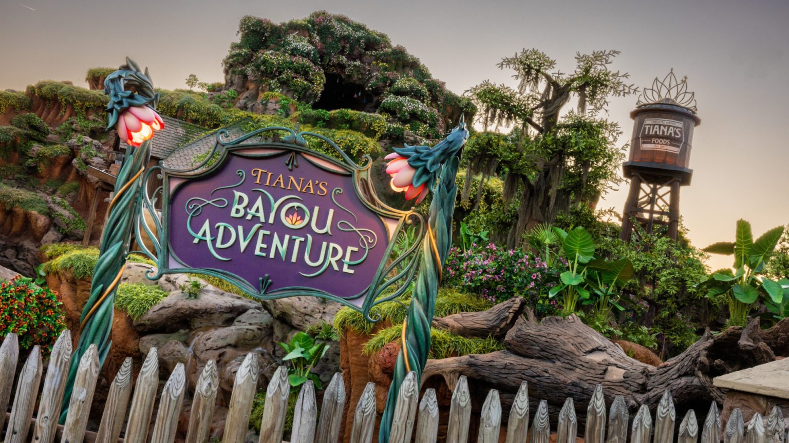 Letreiro de Tiana's Bayou Adventure aparece diante da vegetação e da torre d'água da atração.
