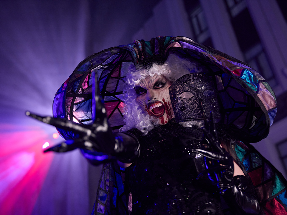 Scareactor com figurino de vampira e gola multicolorida posa em uma scare zone do Halloween Horror Nights.